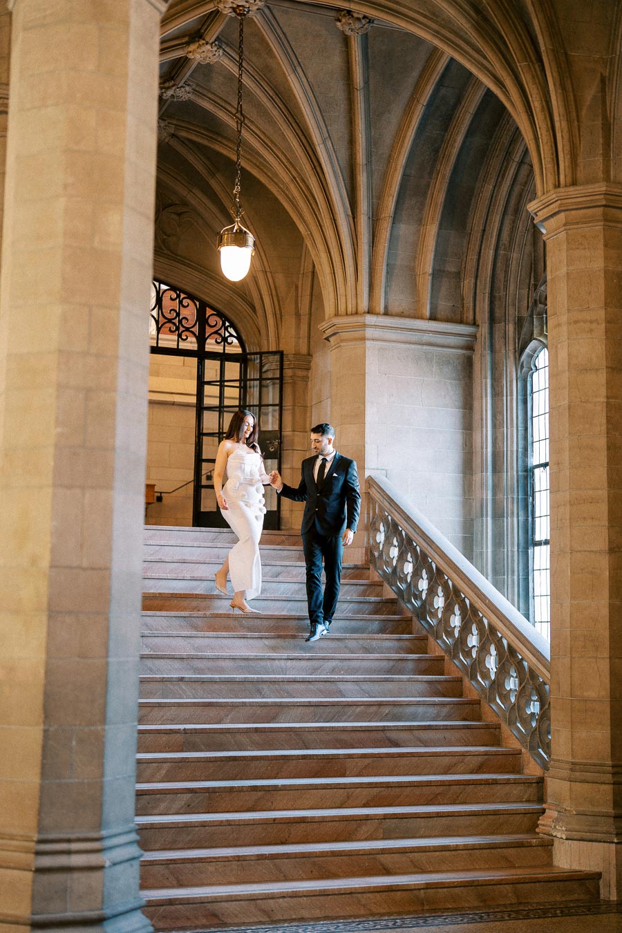 A couple elegantly descending a grand stone staircase in a historic building with ornate arches and large windows.