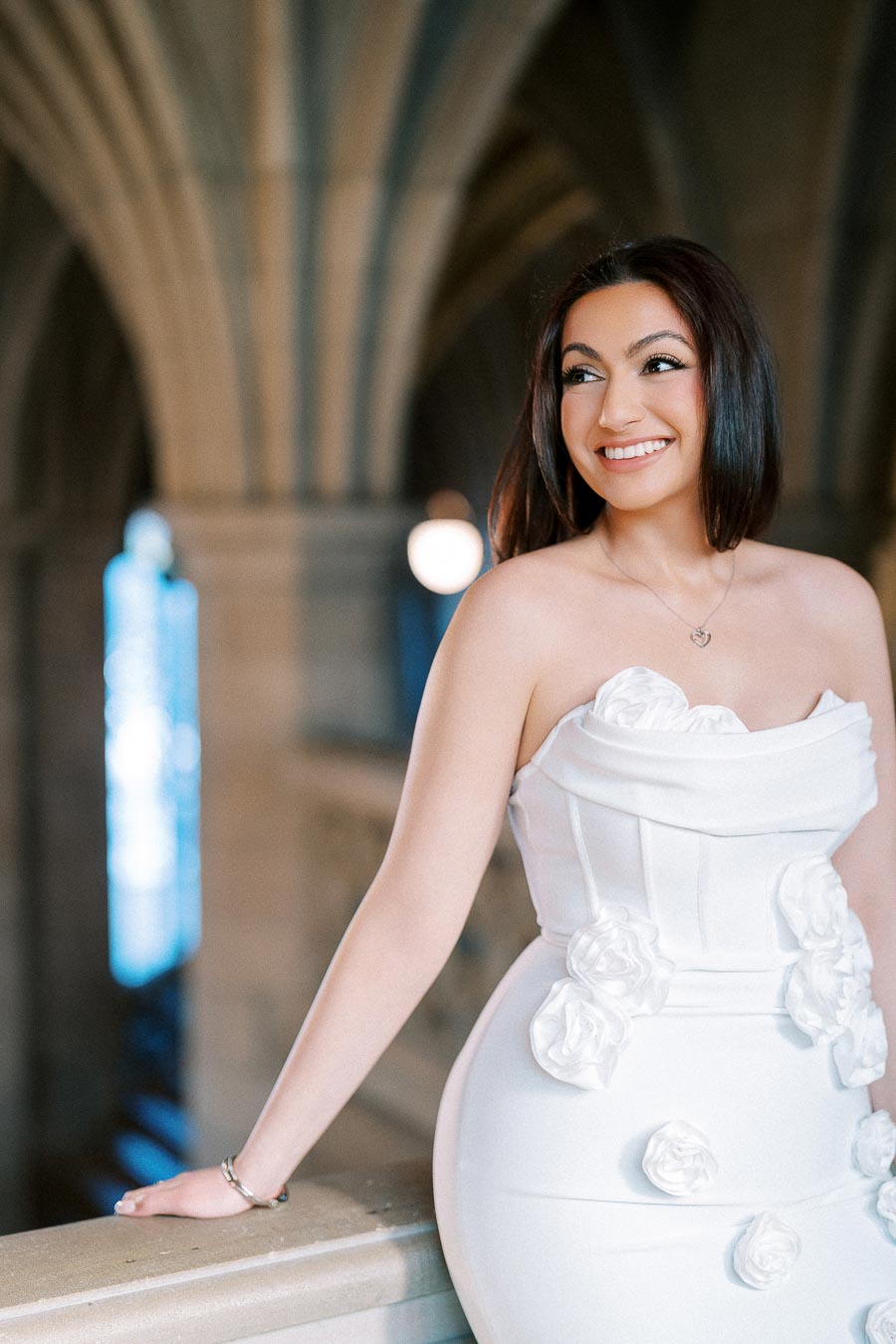 Elegant woman in a white floral dress posing indoors with architectural arches in the background, smiling confidently.