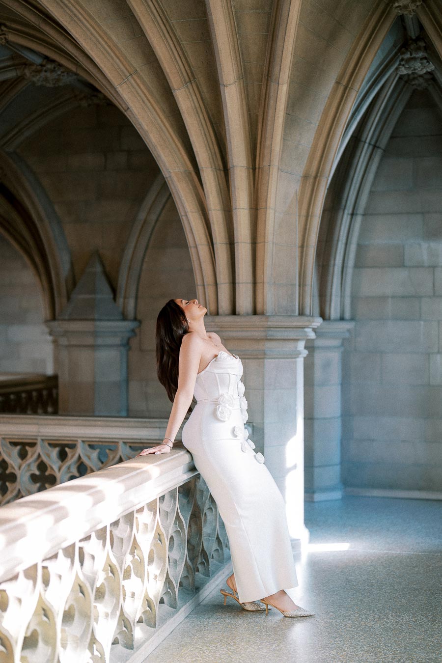 Elegant woman in a white dress leaning against a stone railing in a cathedral-like setting with arched architecture.