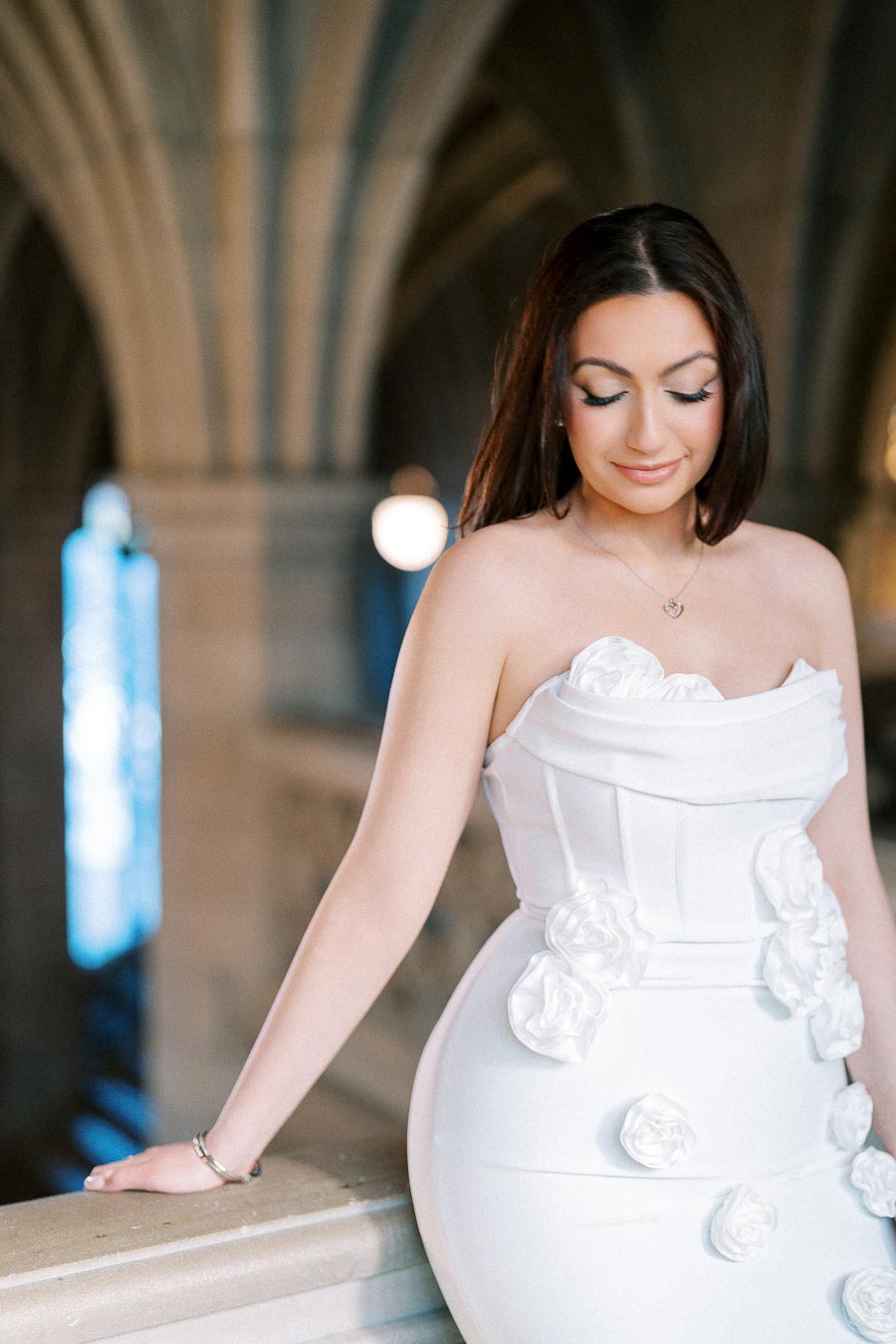 A woman in a white floral dress poses elegantly in a historic building with gothic arches and dim lighting.