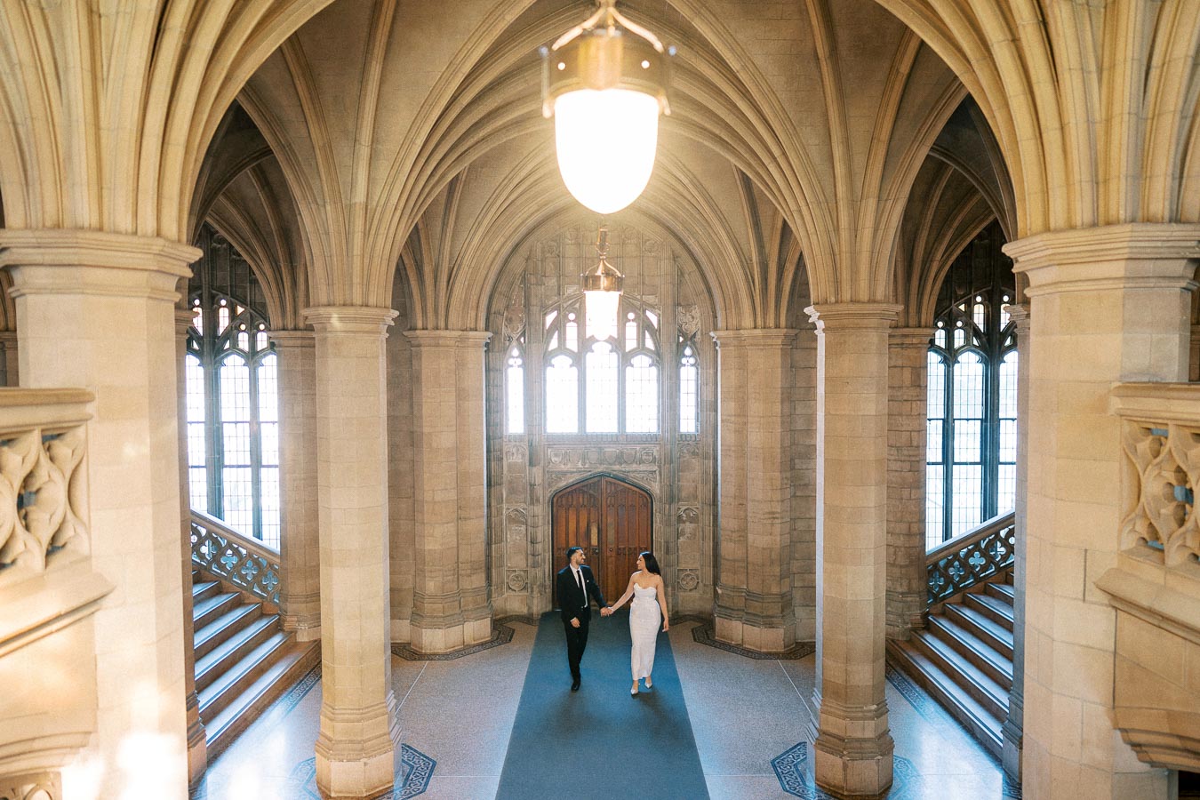 A couple walks hand in hand down a grand, arched hallway with Gothic architecture, featuring ornate columns and decorative