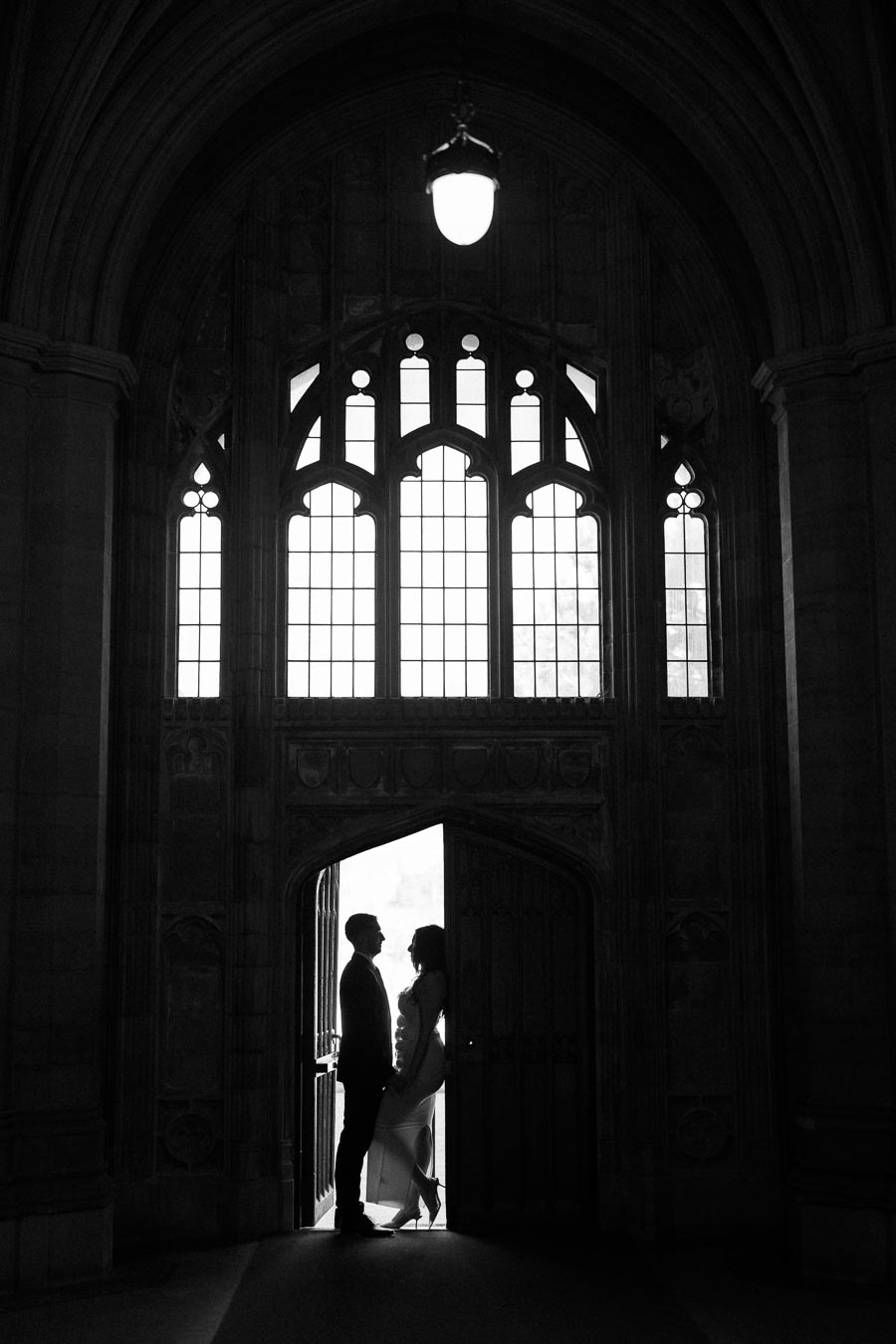 A silhouetted couple stands in an ornate, arched doorway with large Gothic-style windows above, creating a dramatic and