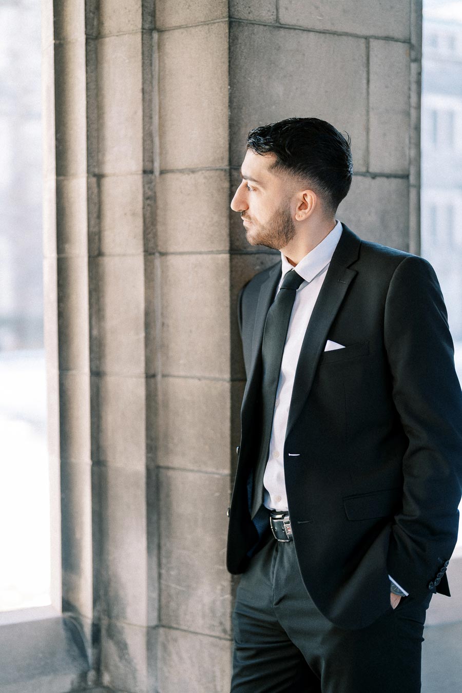 A man in a formal black suit and tie stands thoughtfully against a stone wall, looking out of a window in a well-lit