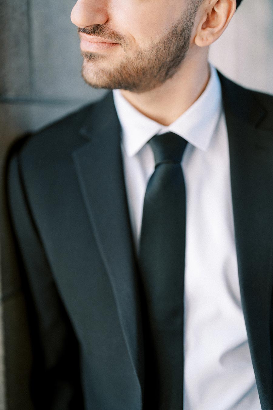 Close-up of a man in a professional black suit and tie, partially facing away, leaning against a wall, showcasing a stylish,