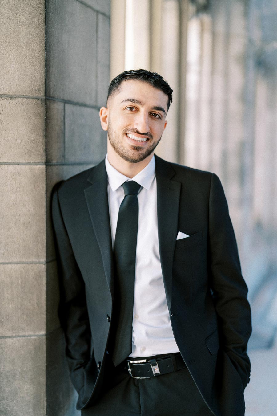 A professional man in a black suit and tie, smiling confidently while leaning against a stone wall.