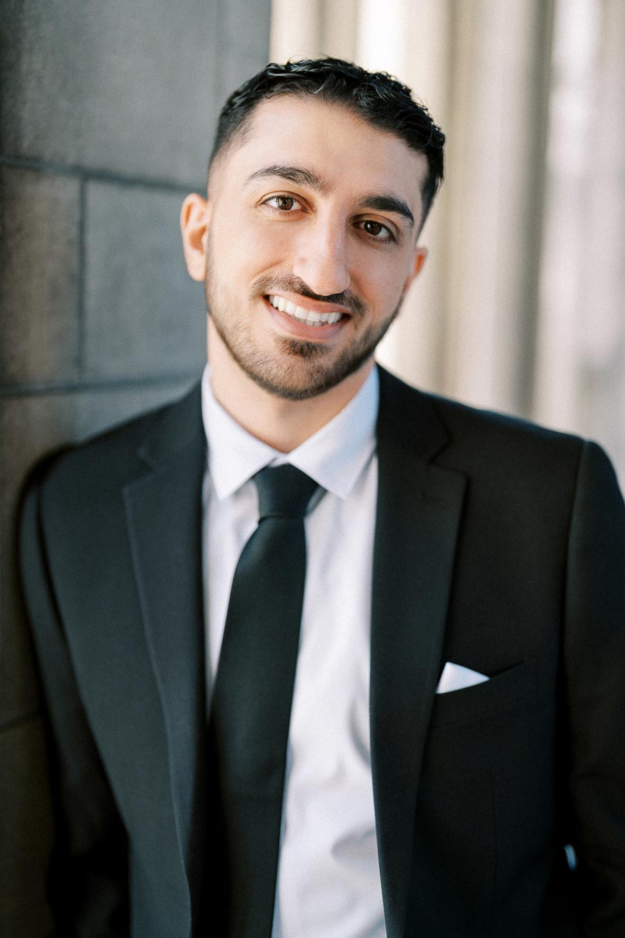 Smartly dressed man in a black suit and tie smiling confidently against a blurred urban background.