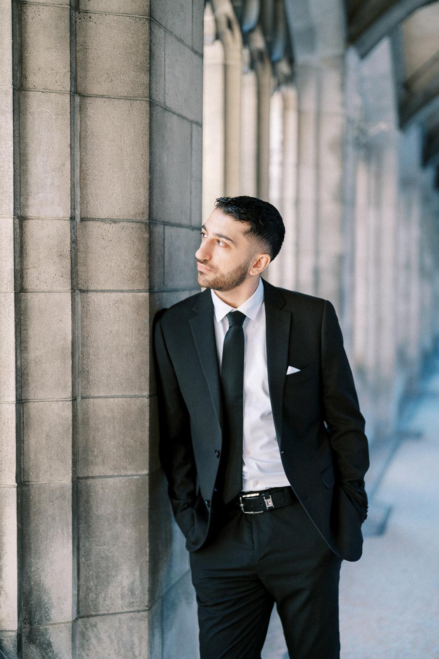 Young man in a black suit leaning against a stone pillar, looking to the side, in an elegant architectural setting.