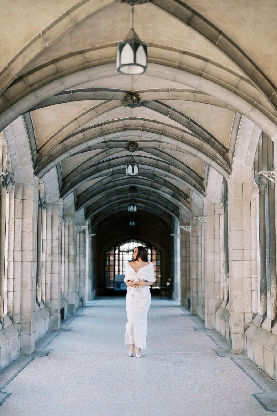 A woman in an elegant white gown walks through a grand stone archway corridor with vaulted ceilings and hanging lanterns.