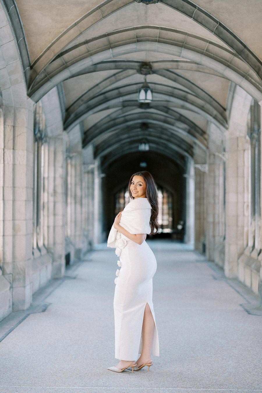 A woman in a white dress poses elegantly in a stone archway corridor, showcasing the architectural details and ornate design