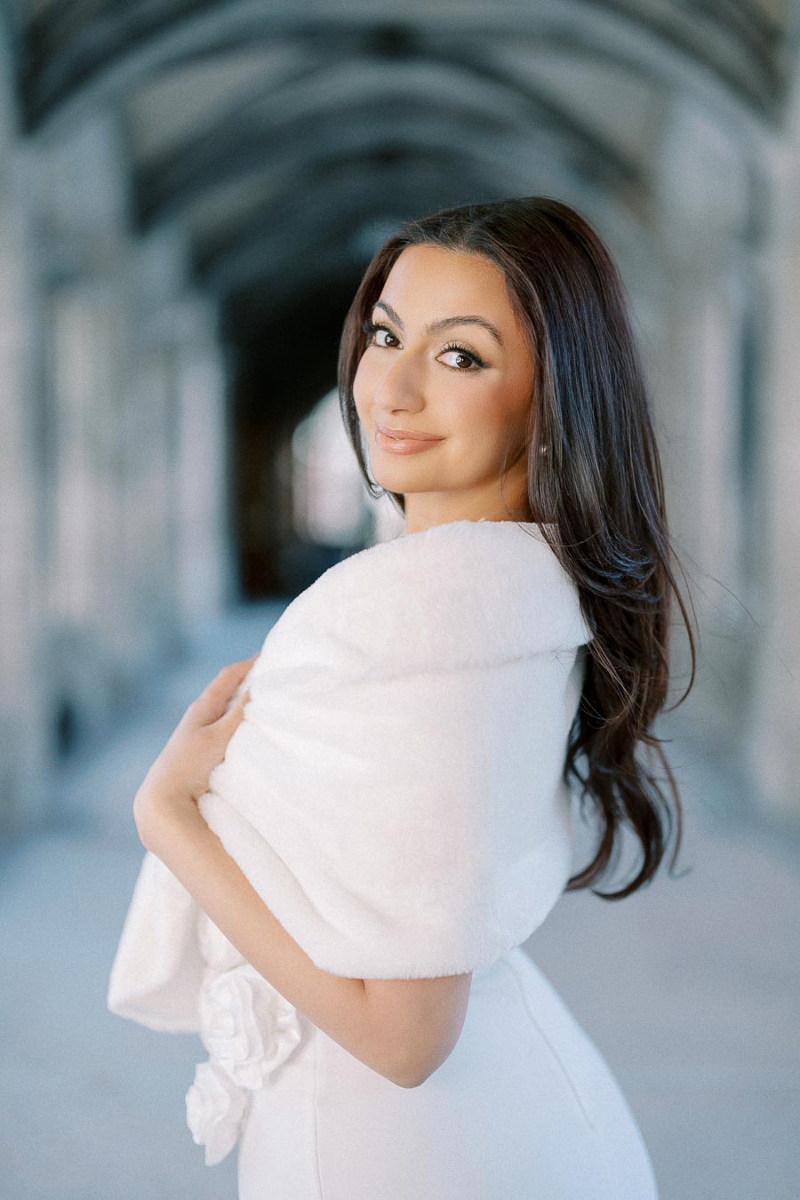Young woman with long brown hair wearing a white shawl, standing in an elegant arched hallway, smiling at the camera.