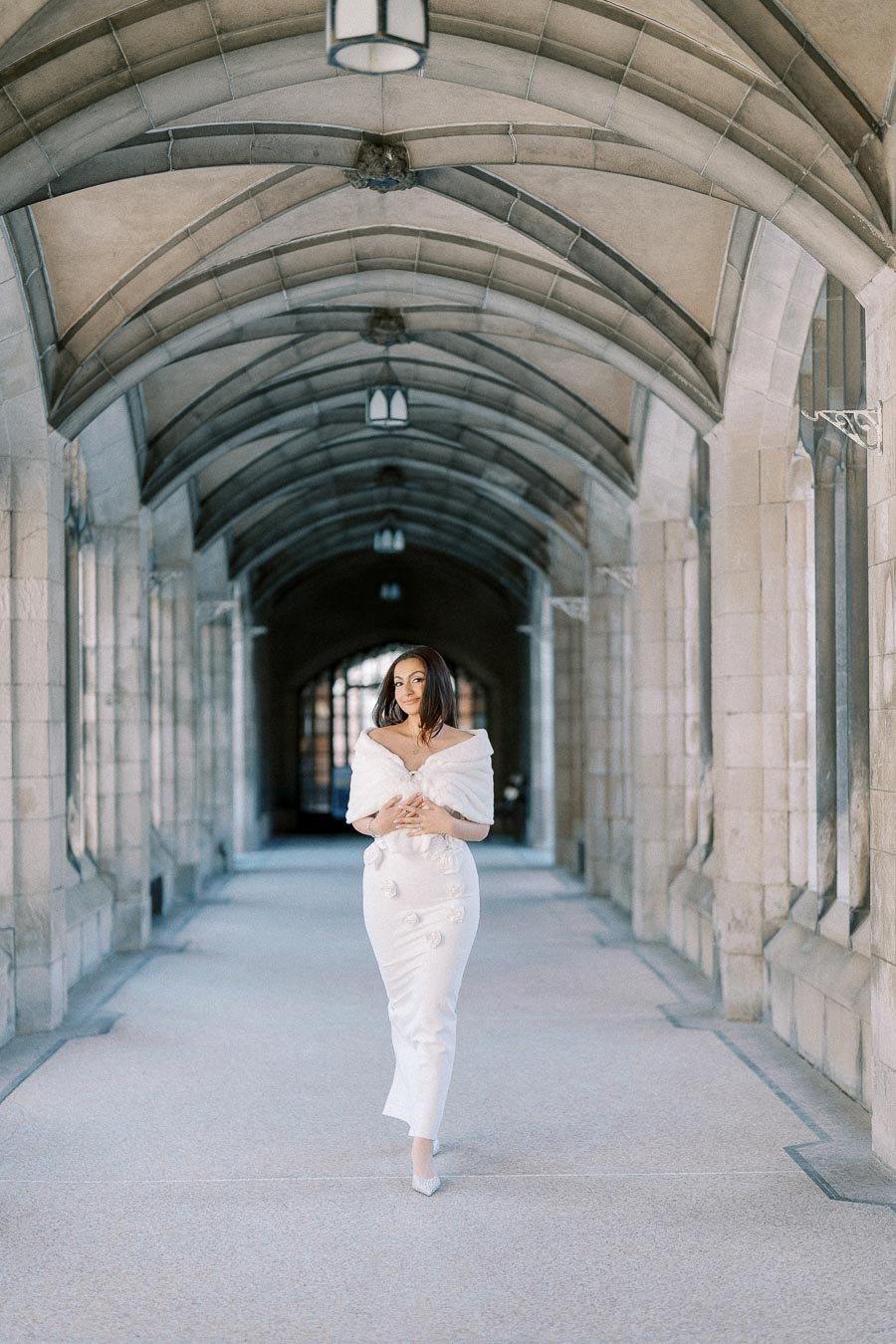 A woman in an elegant white dress and shawl walking through a historic stone archway with vaulted ceilings, conveying a
