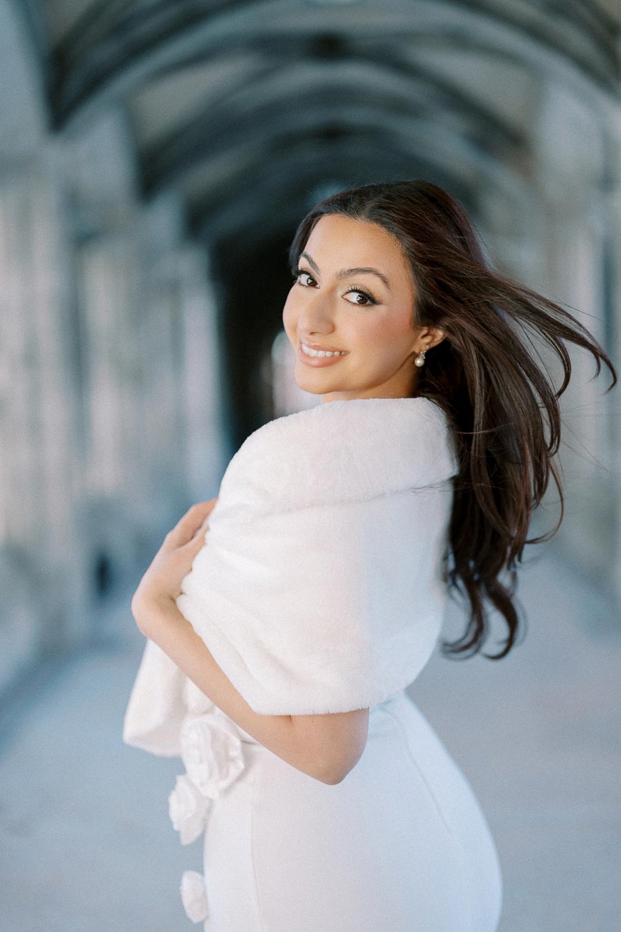 Smiling woman in an elegant white dress and shawl, standing in a beautifully arched stone corridor.