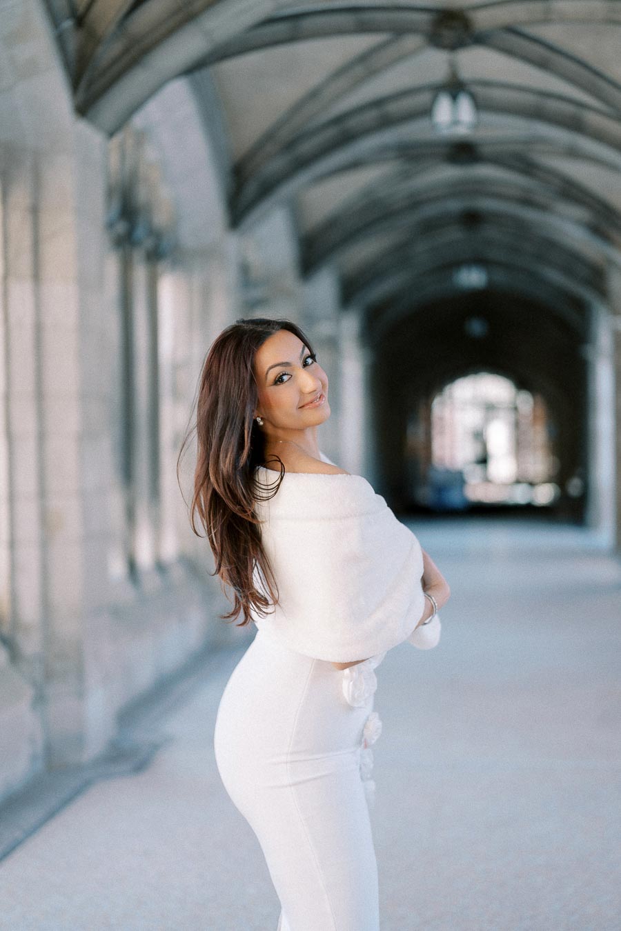 A woman in a white dress standing in a stone archway, looking over her shoulder with a smile.