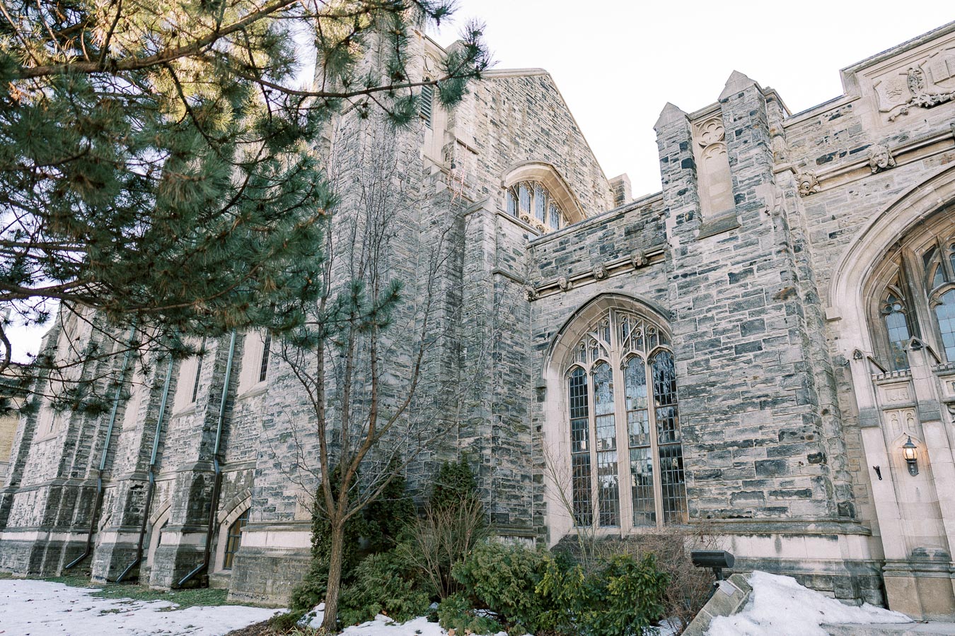 Historic stone building with arched windows, surrounded by evergreen trees and patches of snow, under a clear sky.