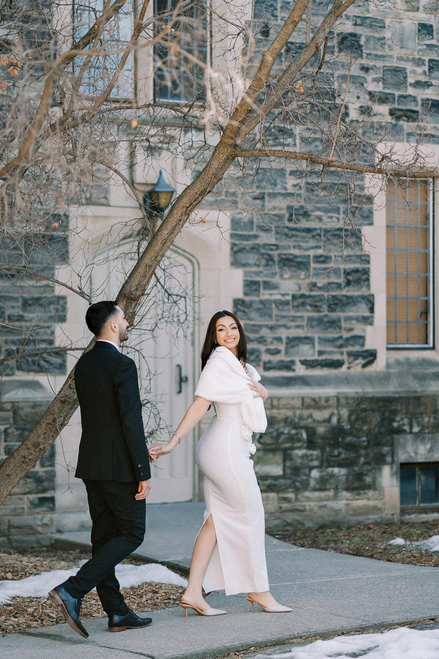 Young couple walking hand in hand past a historic stone building on a winter day, with a woman in a white dress and a man in