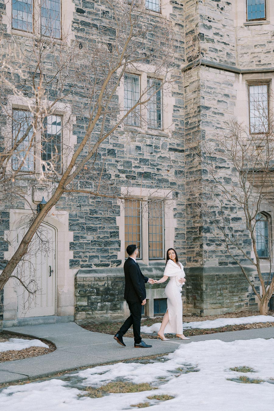 A couple elegantly dressed walking hand-in-hand in front of a historic stone building, surrounded by bare trees and patches