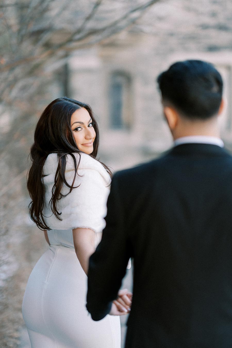 A woman in an elegant white dress and fur shawl looks back with a smile, while holding hands with a man in a black suit,
