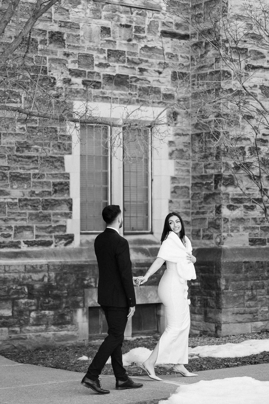 A black and white image of a couple walking hand in hand near a stone building. The woman is smiling and looking back,