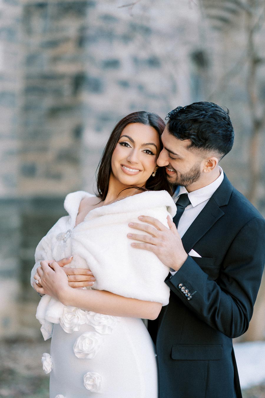 Elegant couple embracing during winter wedding photo shoot, with woman in white fur shawl and man in black suit, smiling