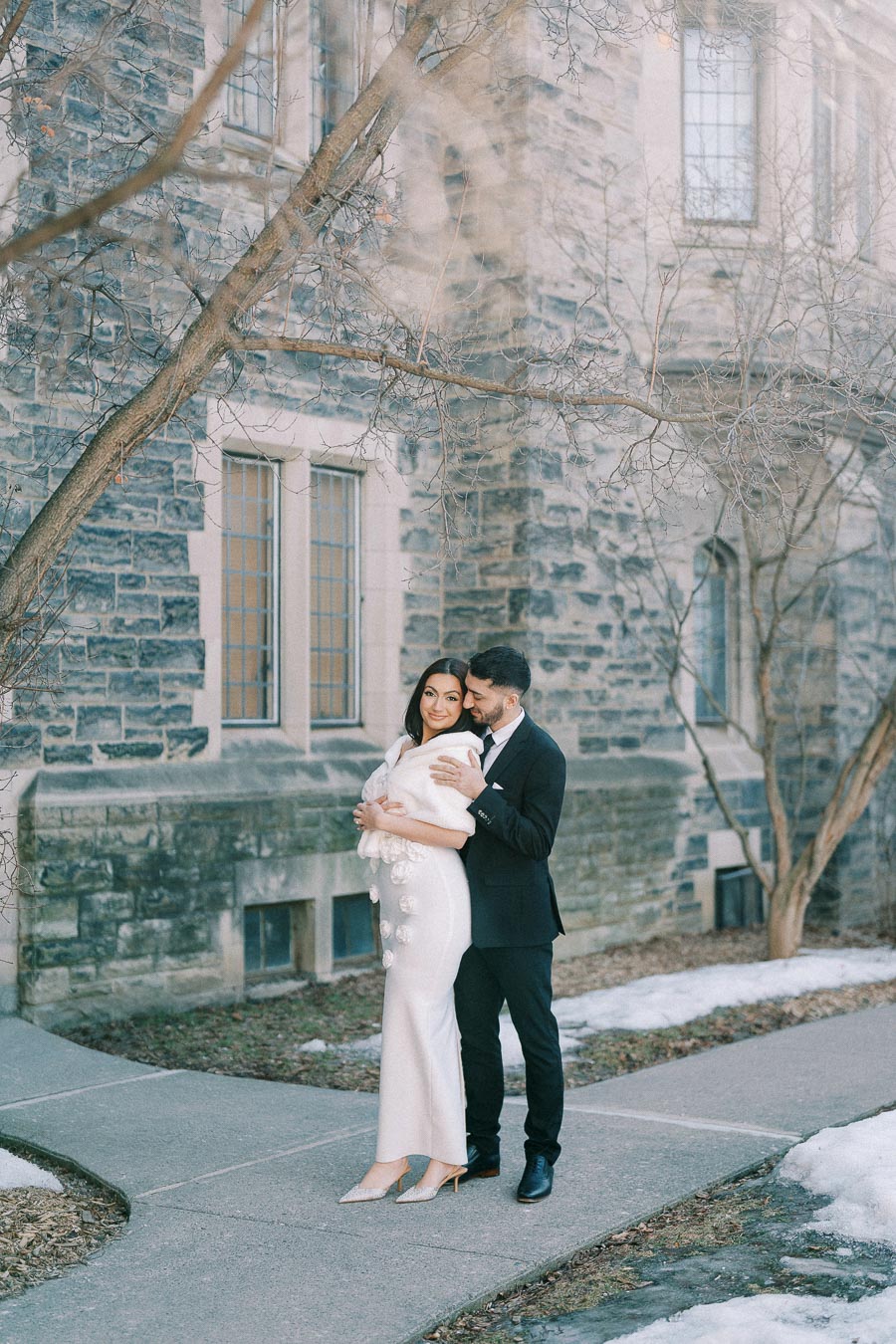 A couple dressed elegantly in formal attire poses outdoors on a snowy day, standing on a sidewalk by a historic stone
