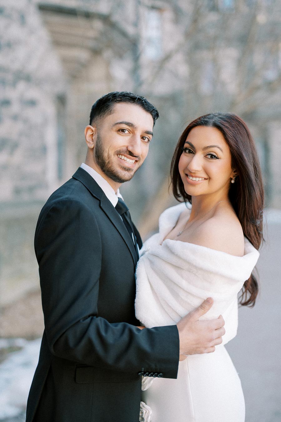 A smiling couple embracing outdoors, dressed in formal attire with a man in a black suit and a woman in a white off-shoulder