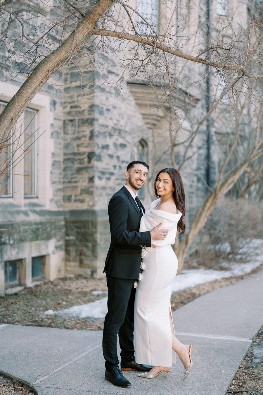A couple posing elegantly in formal attire; the man in a black suit and the woman in an off-the-shoulder white gown, stand