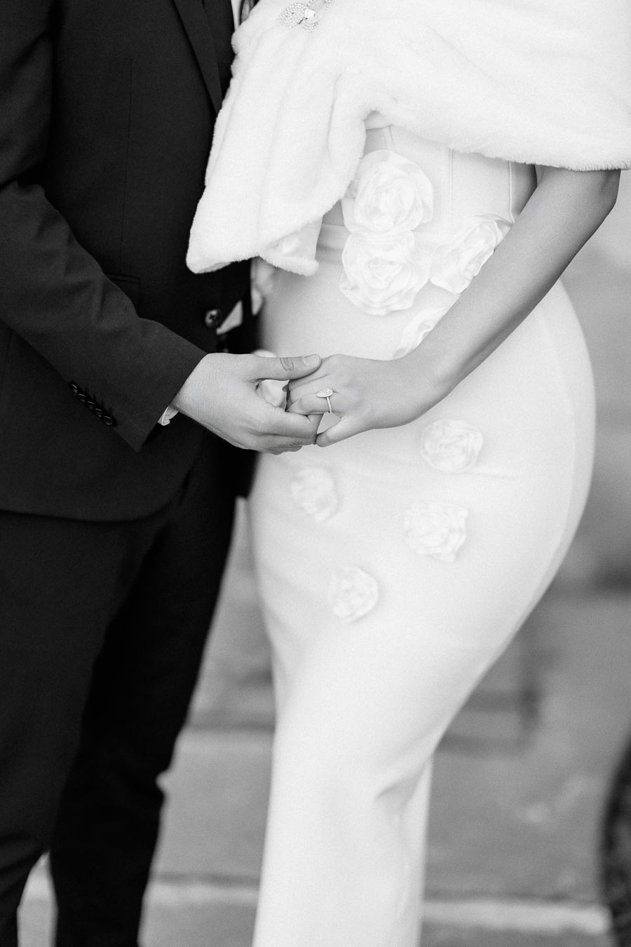 Elegant black and white photo of a couple holding hands, showcasing a bride in a floral detailed dress and fur shawl,
