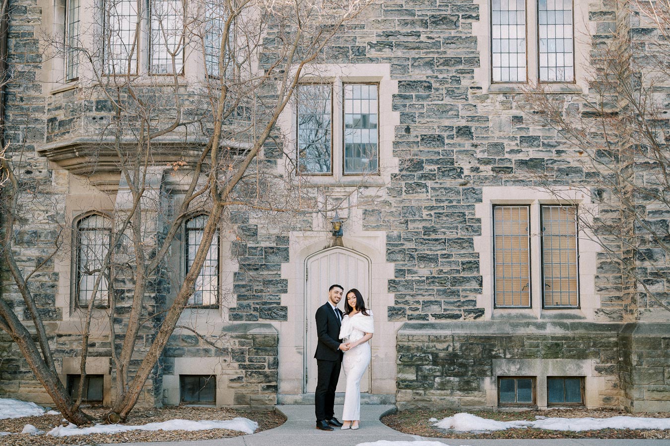 A couple posing in front of a historic stone building with arched windows and a classic wooden door, surrounded by early