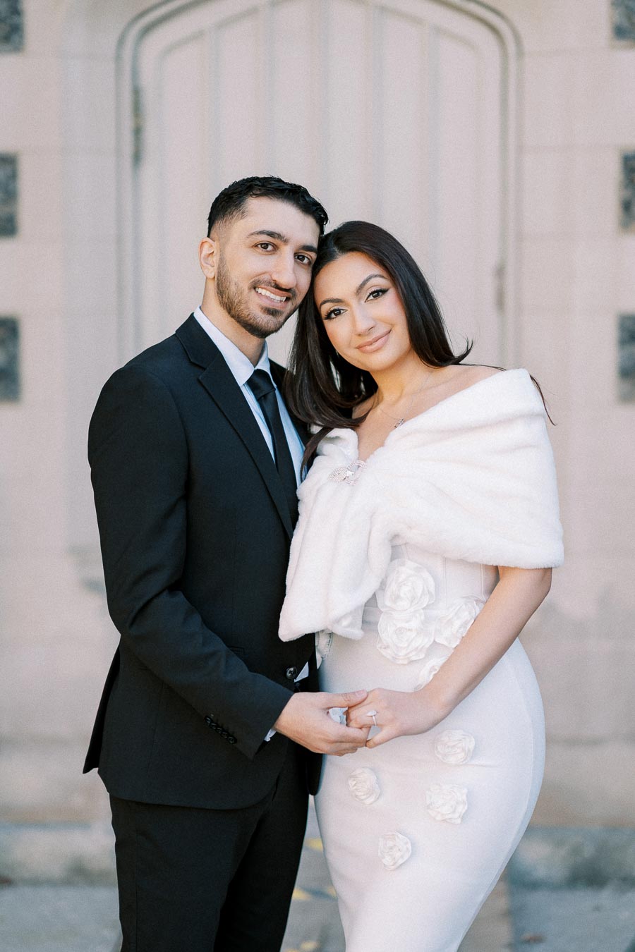 Elegant couple in formal attire posing for engagement photo; man in black suit and woman in white dress with floral details,