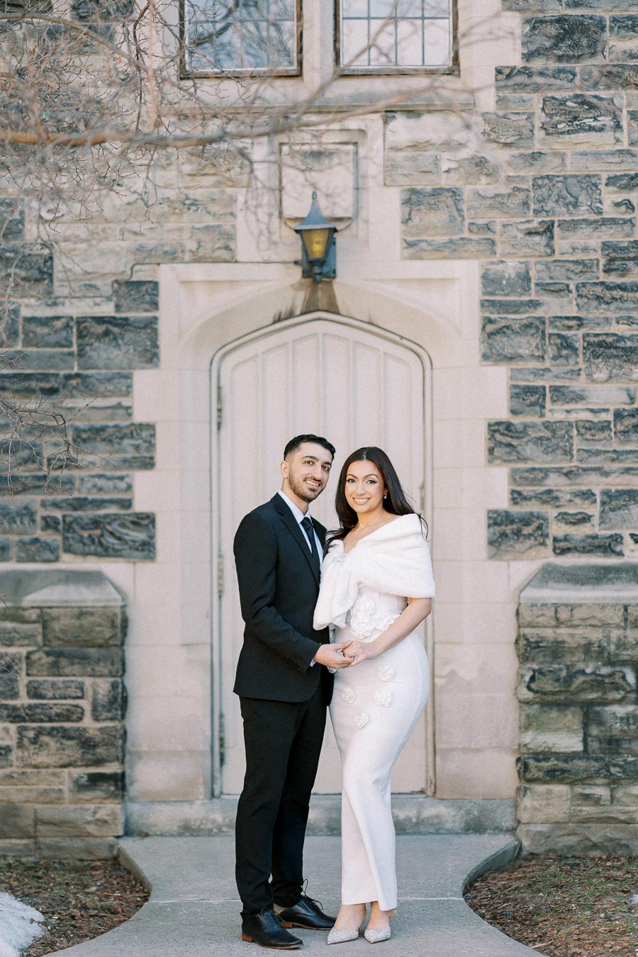 Elegant couple posing in front of a historic stone building, with the woman in a white dress and the man in a black suit,