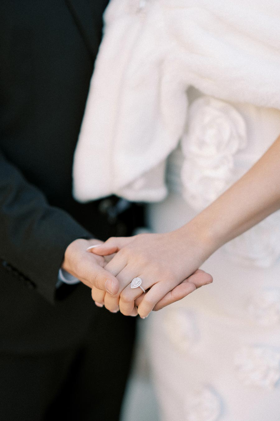 A close-up image of a couple holding hands, showcasing the bride's elegant diamond engagement ring, with a backdrop of her