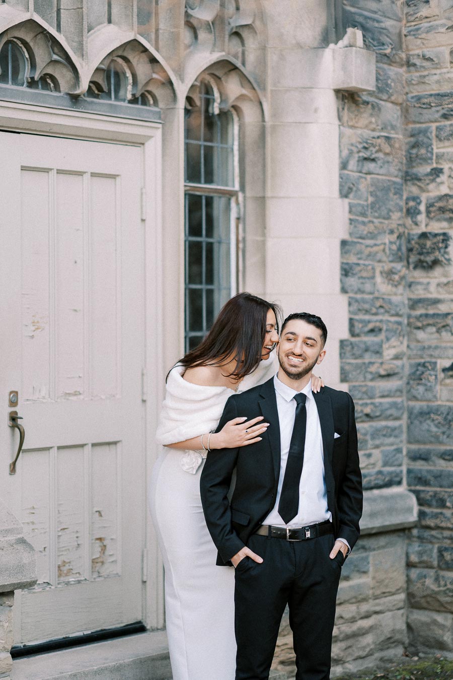 A couple in formal attire posing happily near a historic stone building with gothic arch windows and a wooden door.