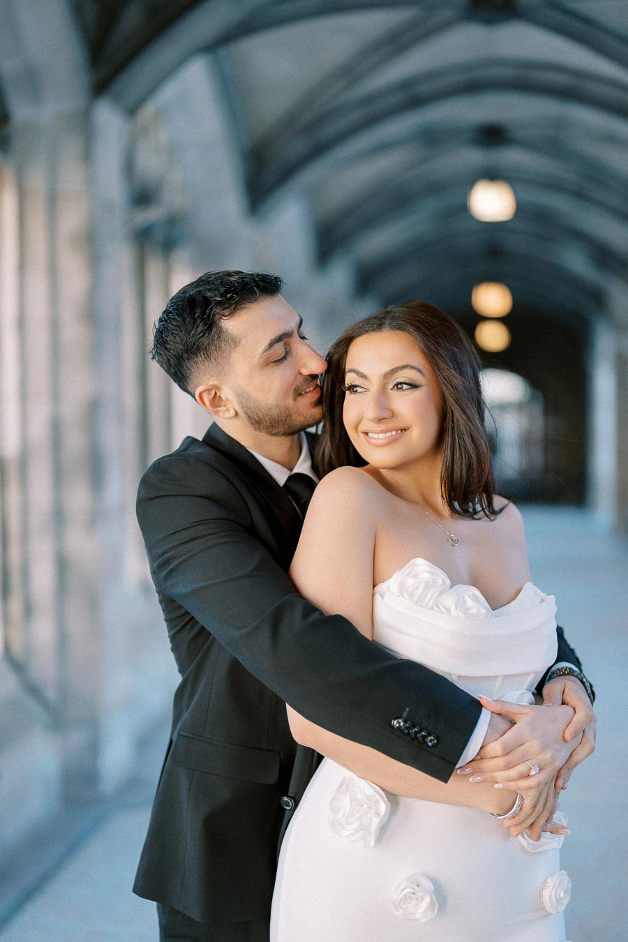 A couple in formal attire embraces in a romantic, arched hallway setting; the man in a black suit and tie, and the woman in