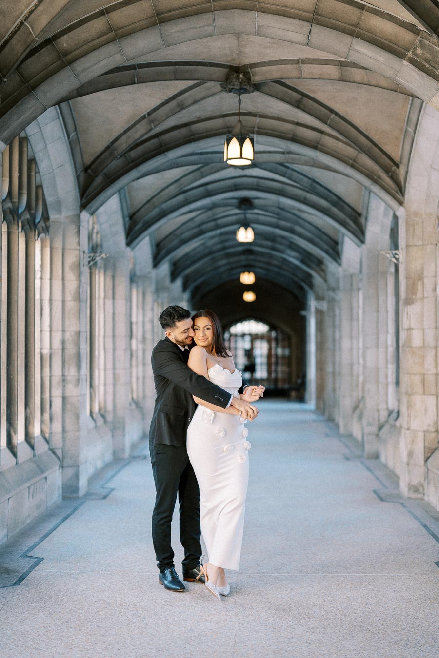 A couple embraces in a decorative stone archway, with the man wearing a black suit and the woman in a white dress adorned