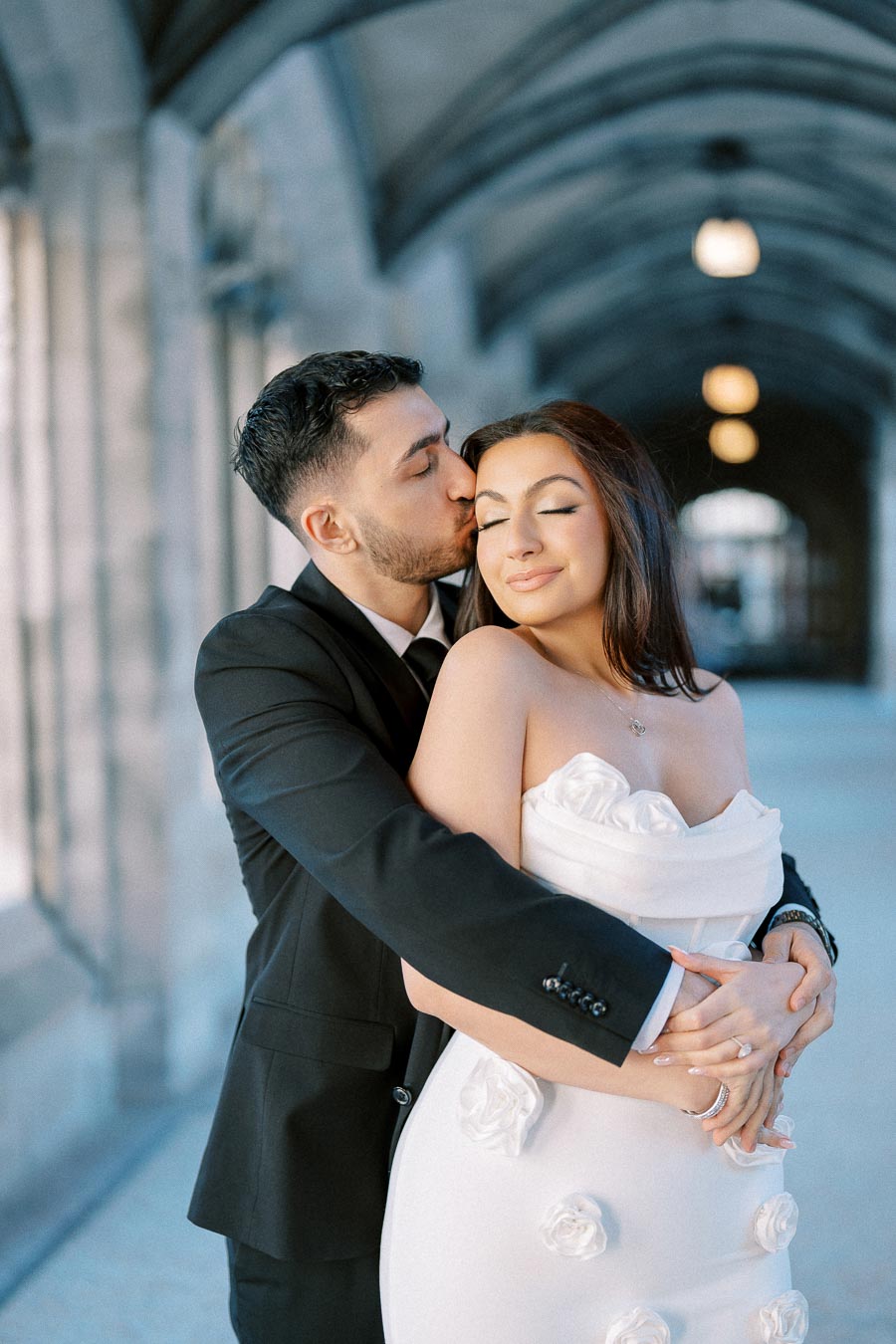 A romantic couple embraces under an elegant arched hallway. The man in a black suit kisses the woman in a white strapless