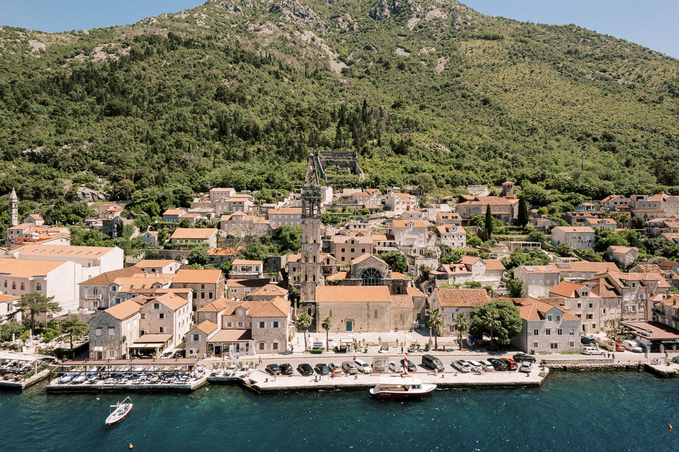 Aerial view of a quaint coastal town with terracotta roofs nestled against a lush green hillside, featuring a historic church tower and a serene waterfront, perfect for travel and tourism enthusiasts.