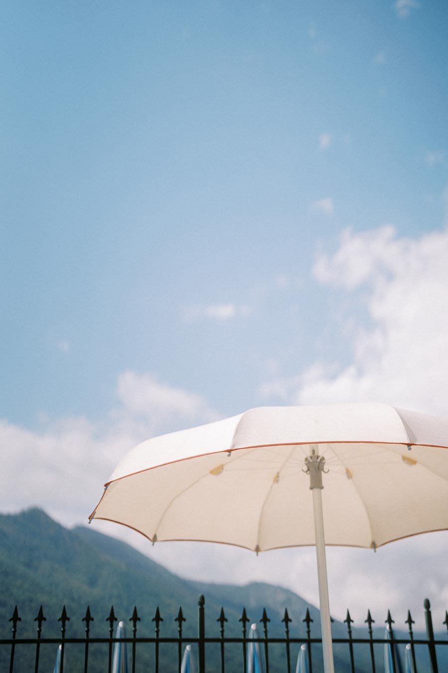 White umbrella under clear blue sky with mountain backdrop and iron fence in foreground, symbolizing summer relaxation and outdoor leisure.