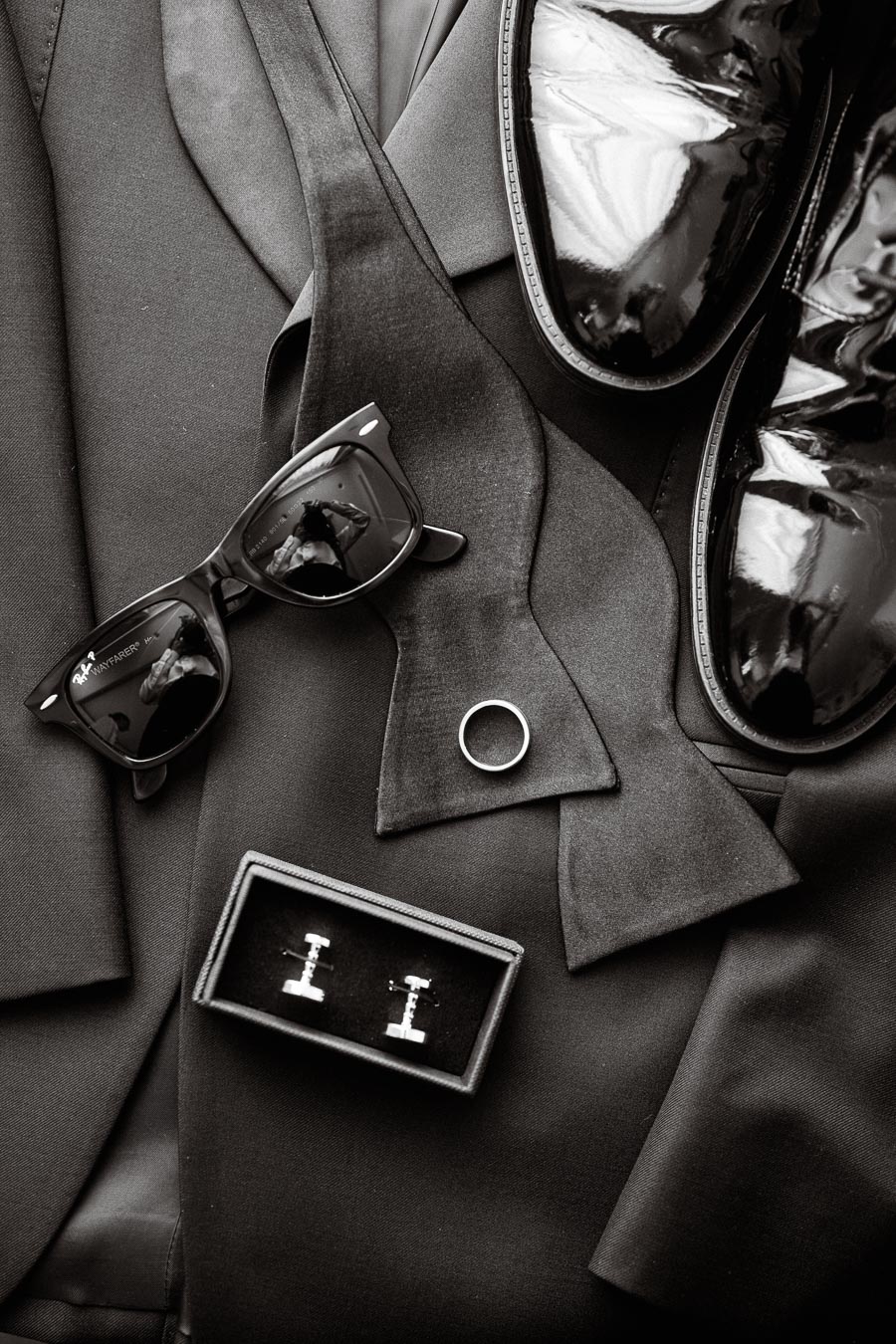 Elegant men's accessories including polished dress shoes, a bow tie, sunglasses, a ring, and cufflinks on a black suit, symbolizing sophistication and style.