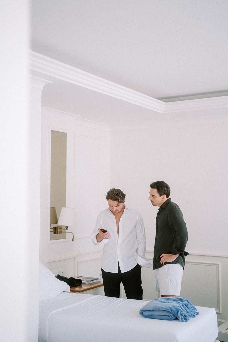 Two men in a modern hotel room, one in a white shirt looking at a phone, the other in a dark shirt watching. A neatly made bed with a folded blue blanket in the foreground. Bright, minimalist interior design.
