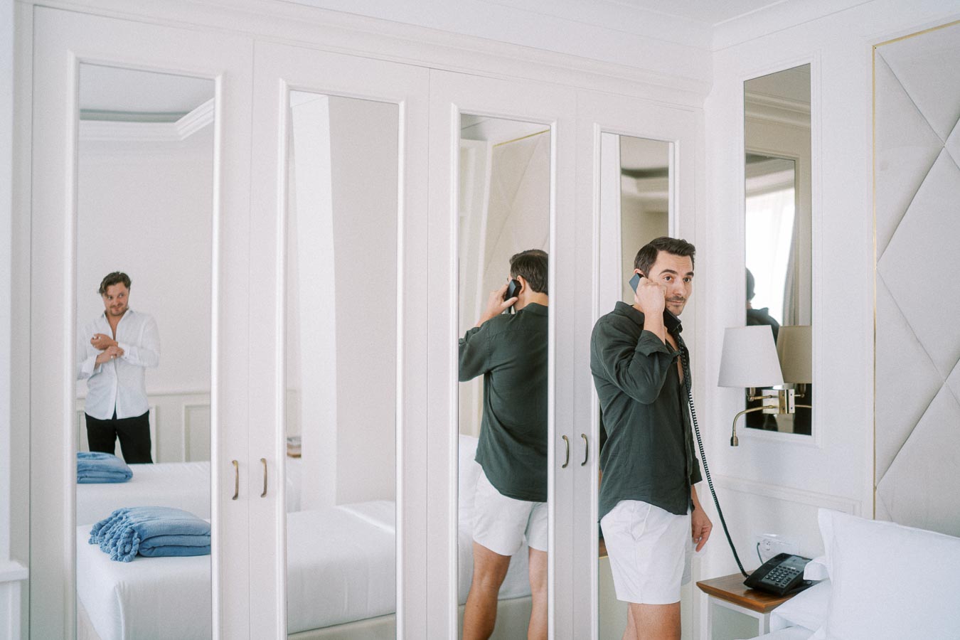 Man standing in a stylish hotel room, talking on a phone with a reflection in mirrored closet doors. Another man buttoning his shirt is visible in the room. The space is bright, featuring modern decor and neatly arranged linens.