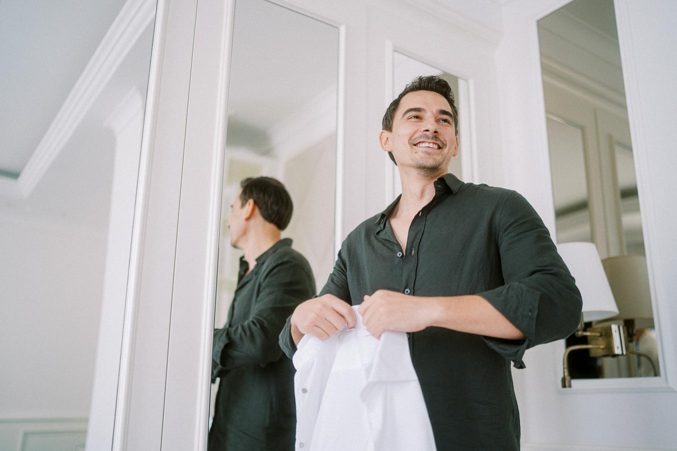 Man smiling while holding a white shirt in a brightly lit room, standing in front of mirrored wardrobes.