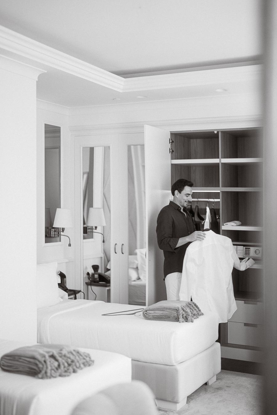 Man selecting a white shirt from a hotel room wardrobe, showcasing a neatly organized and luxurious accommodation setting with white interior decor and modern amenities.