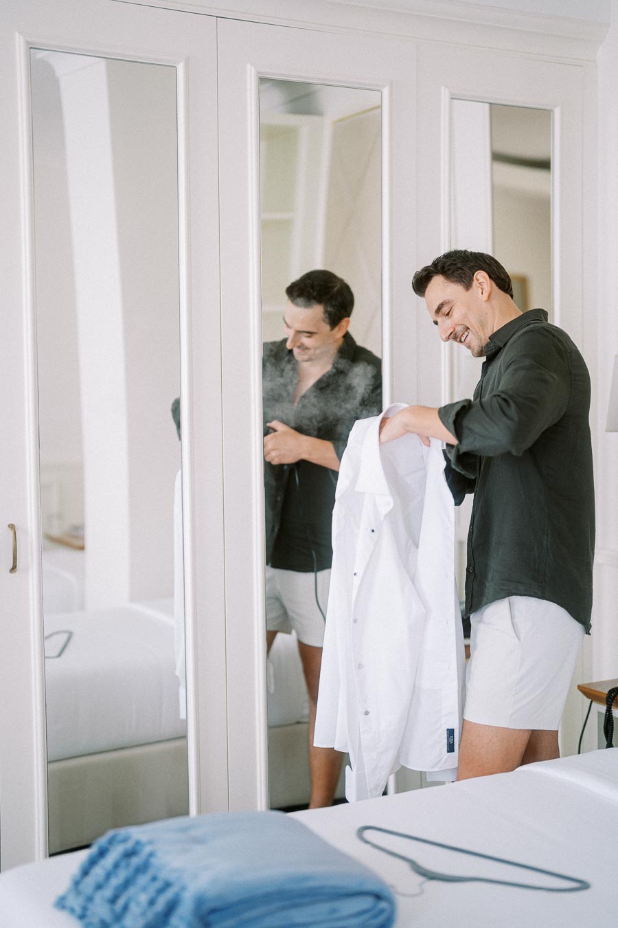 Man steaming a white shirt in front of a mirror, wearing casual clothes in a bright, modern bedroom.