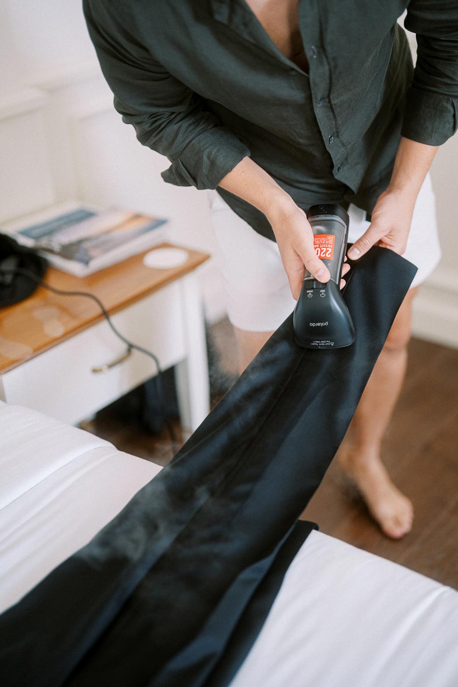 A person in a dark shirt and shorts using a garment steamer to remove wrinkles from a black piece of clothing on a bed, with a nightstand in the background.