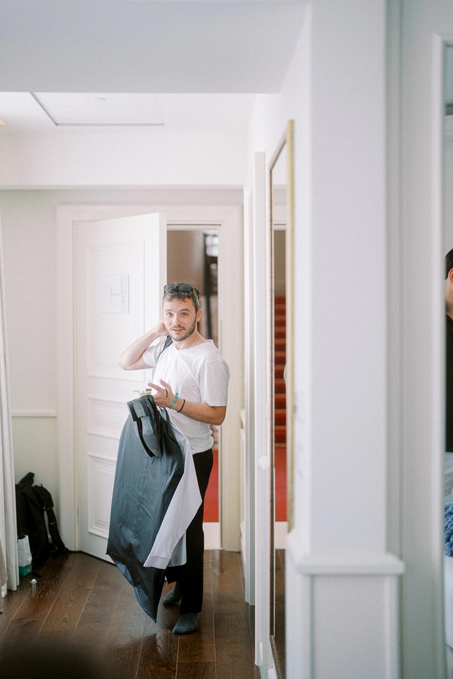 Man in a casual white t-shirt holding a garment bag while standing in a bright, modern room with hardwood floors and open doors, suggesting a preparation moment.