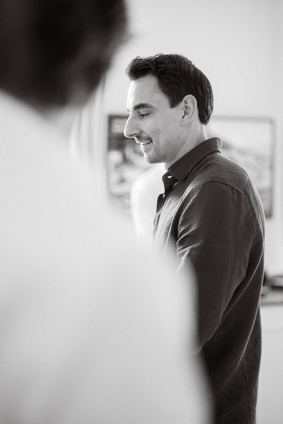 Black and white portrait of a smiling man in a casual shirt, standing indoors with soft focus in the foreground, conveying a relaxed and friendly atmosphere.