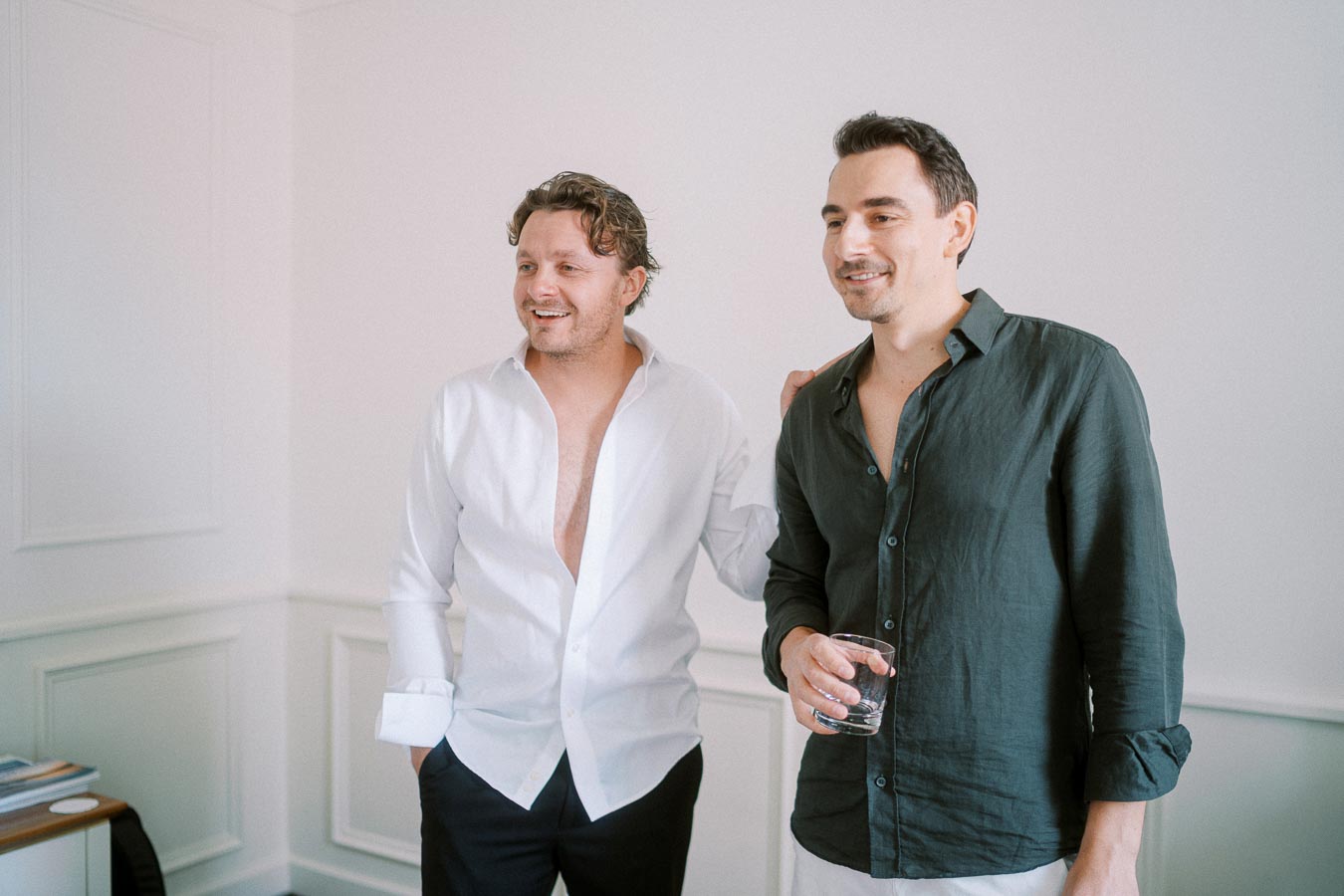 Two men standing together in a brightly lit room; one in a white shirt and the other in a dark green shirt holding a glass.