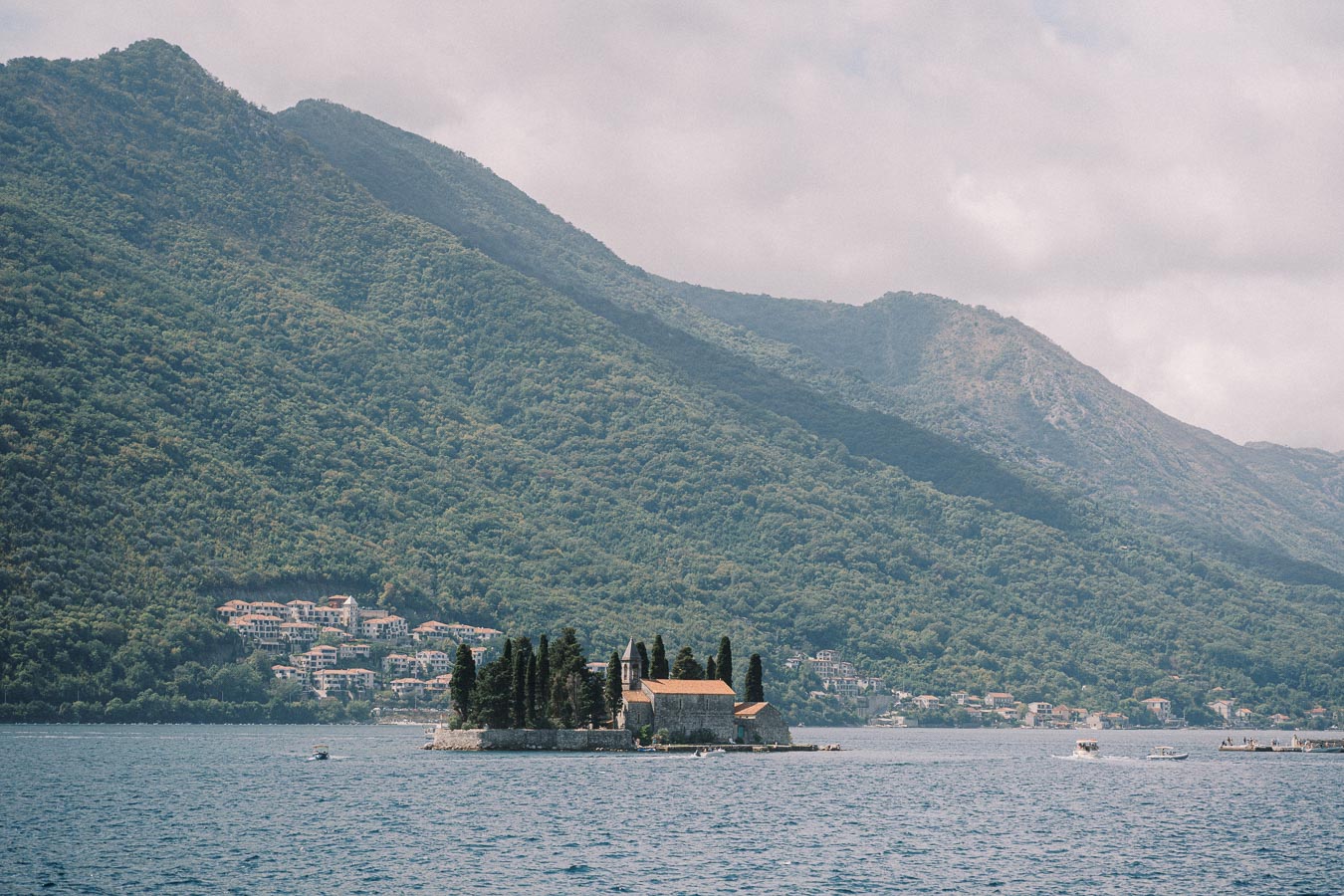 A scenic view of a small island with a historic church surrounded by tall cypress trees, set against a backdrop of lush green mountains and calm blue waters.