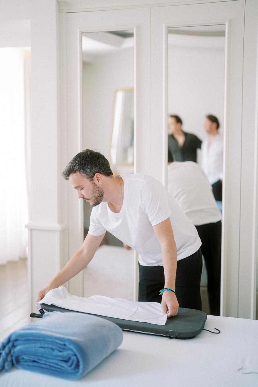 Man in white t-shirt packing a garment bag in a bright hotel room with mirrors.