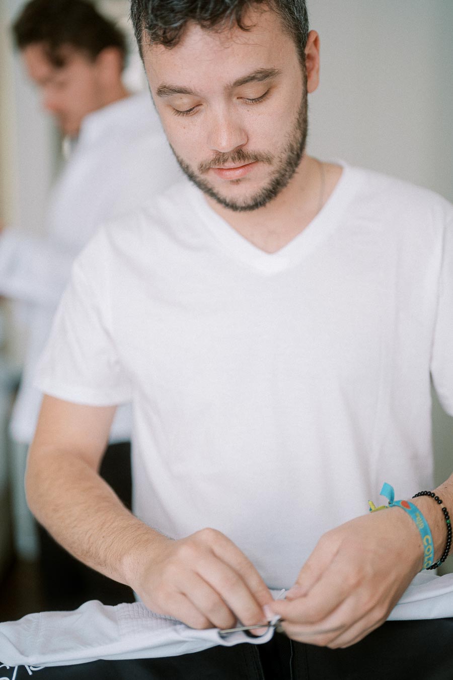 Man in casual white t-shirt concentrating on folding clothes neatly, with a wristband on one arm and another person out of focus in the background, in a bright indoor setting.